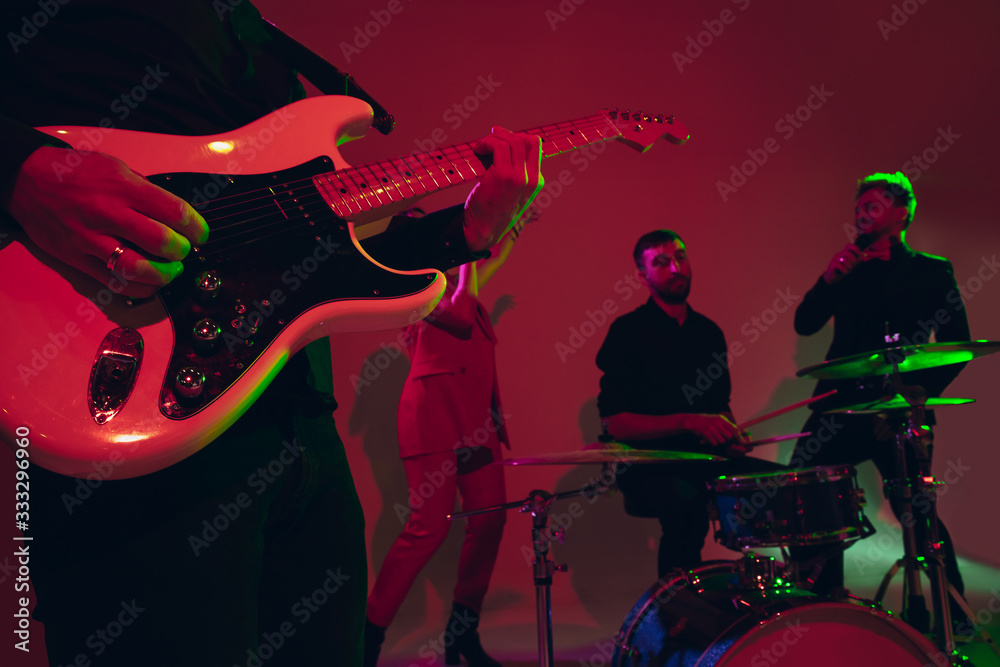 Obraz premium Young and joyful caucasian musicians performing on red studio background in neon light. Concept of music, hobby, festival. Colorful portrait of modern artist. Attented and inspired. Art, cover band.