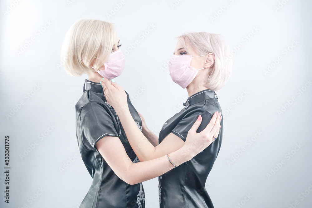 two European-looking women are dressed in black Chinese dresses and medical face masks. blonde girls do not keep a safe distance during coronovirus covid-19. females in white photo Studio. two European-looking women are dressed in black Chinese dresses and medical face masks. blonde girls do not keep a safe distance during coronovirus covid-19. females in white photo Studio.