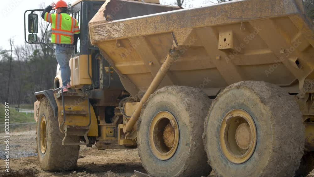 Dump truck driver climbs up the stairs to the cab and points out over the construction project. Dump truck driver climbs up the stairs to the cab and points out over the construction project.