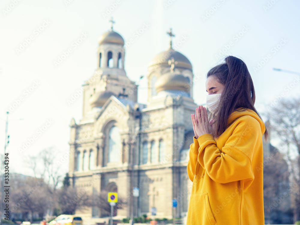 young woman wearing protective mask praying outside near church