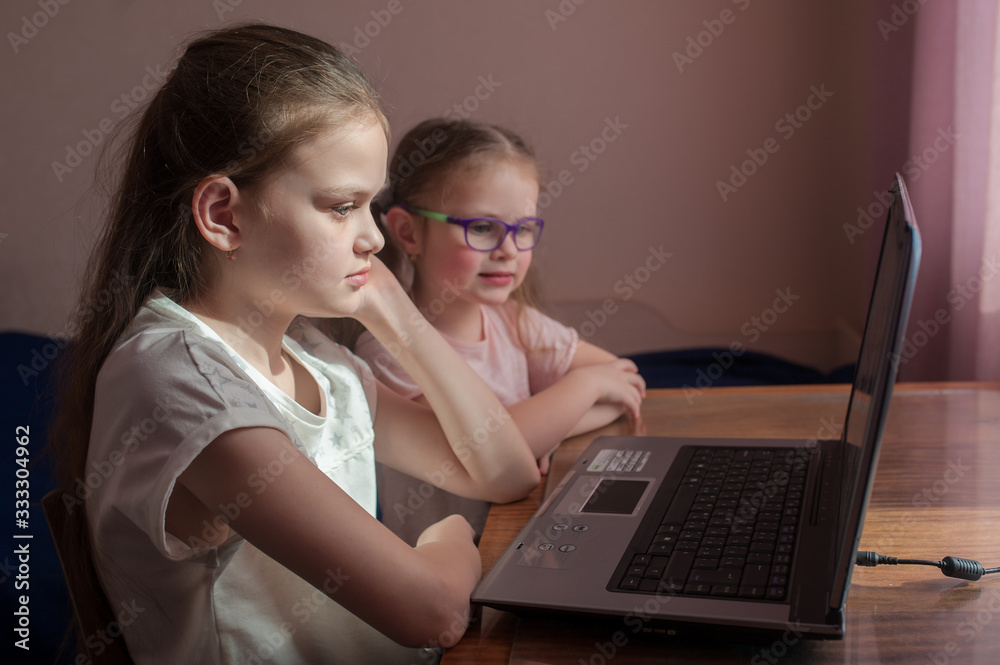Two girls, the oldest and the youngest, are engaged at a table on a laptop. Little sister with glasses. Two girls, the oldest and the youngest, are engaged at a table on a laptop. Little sister with glasses.