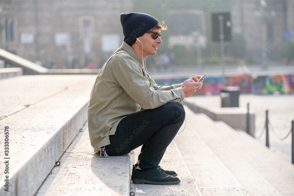 Portrait of young smiling man isolated on outside outdoor background uses a cellphone, earphones Portrait of young smiling man isolated on outside outdoor background uses a cellphone, earphones