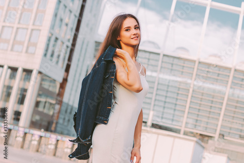 Portrait of young fashionable cute woman wearing stylish denim jacket and dress walking city streets and smiling. Attractive happy trendy hipster girl posing outdoors. Modern city fashion and style