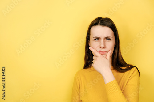 Frowning woman wearing sweater holding hand on chin, purses lips with clueless expressions, looking at camera feels distrustful studio head shot portrait isolated on yellow background