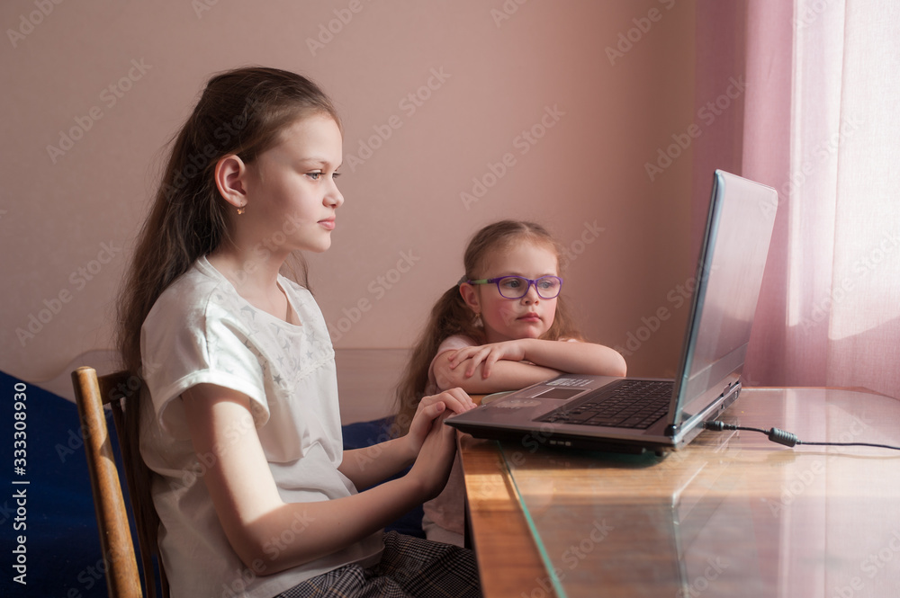Two girls, the oldest and the youngest, are engaged at a table on a laptop. Little sister with glasses. Two girls, the oldest and the youngest, are engaged at a table on a laptop. Little sister with glasses.