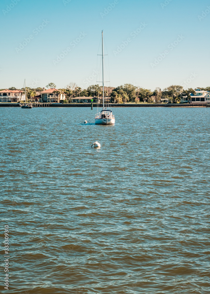 Obraz premium Small Sailboat Lying At Anchor with Houses Along the Coast Behind