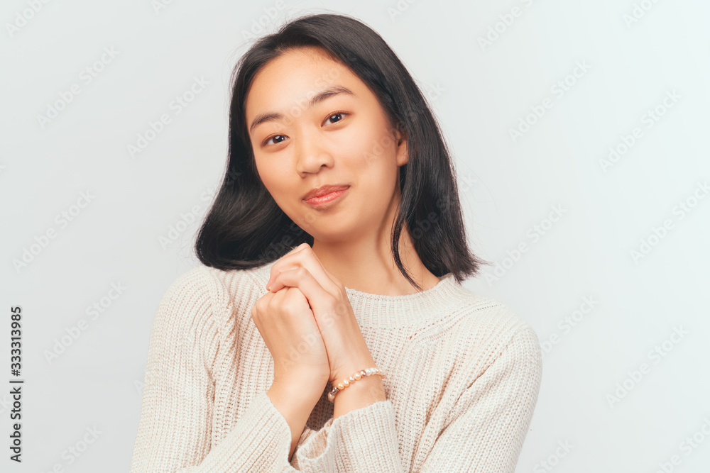 Delighted girl presses hands to face looking at camera. Beautiful young woman Asian appearance with black hair and brown eyes dressed in knitted warm sweater stands isolated white background in Studio Delighted girl presses hands to face looking at camera. Beautiful young woman Asian appearance with black hair and brown eyes dressed in knitted warm sweater stands isolated white background in Studio