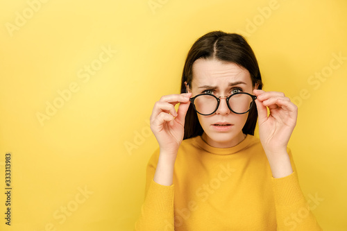 Emotion young woman keeps hands on rim of spectacles looks with omg expression gasps from shock, hears astonishing unbelievable news, dressed in sweater, isolated on yellow background with copy space