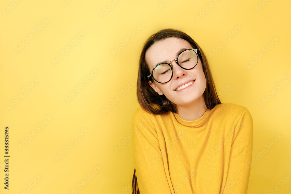 Lovely merry woman in eyeglasses has toothy smile, closes eyes, feels pleasure from good compliment, wears jumper, stands against yellow background with copy space for your advertisement