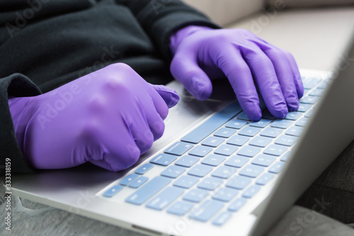 Person in protective purple medical rubber gloves sits on grey sofa at home and works or studies on laptop during quarantine. Remote work during coronavirus pandemic.