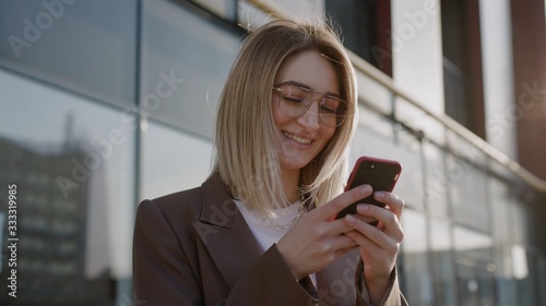 close up view of young attractive woman near business building smile using phone walking at break in the city  professional female employer typing text message on cellphone spring