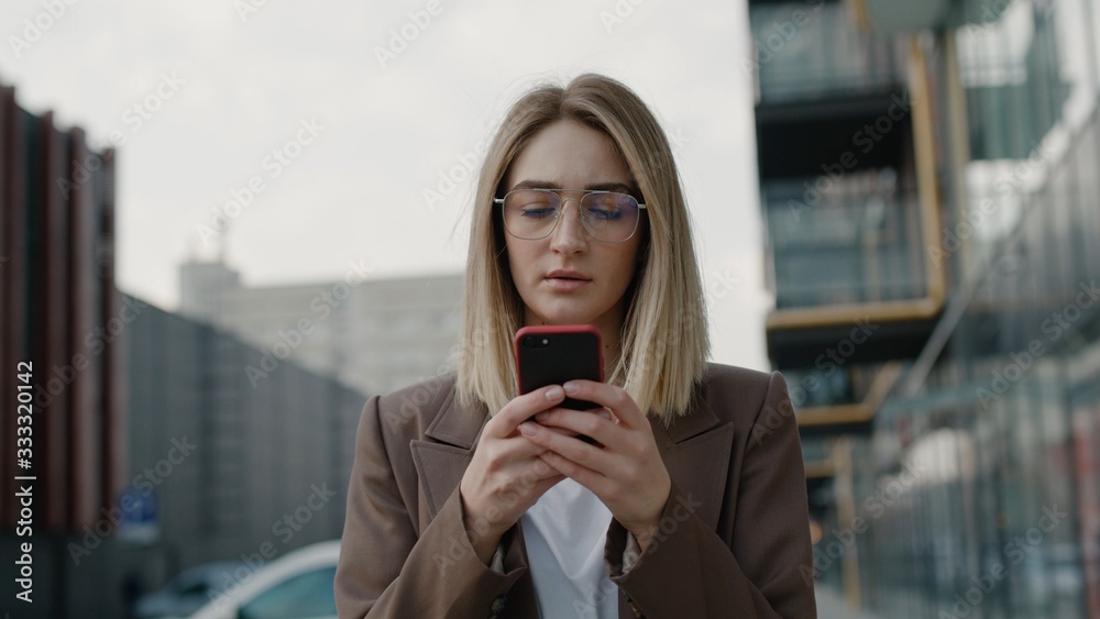Fototapeta premium Portrait pretty young businesswoman using smartphone at street Serious woman typing message on smartphone outdoor. European girl holding mobile phone in hand outdoor. Business woman chatting online