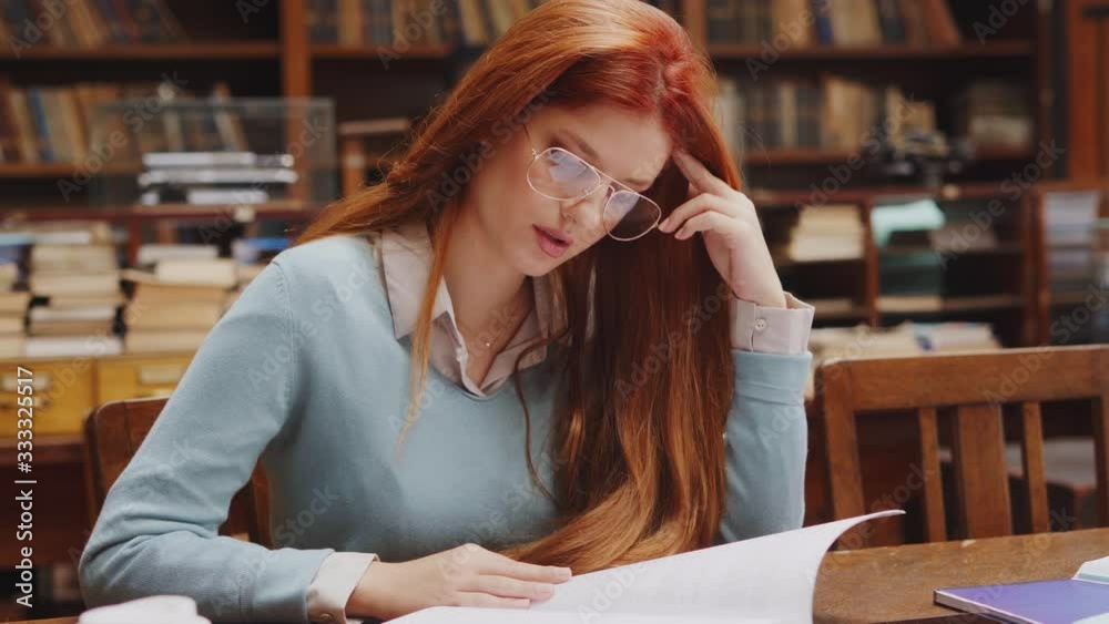 Woman with spectacles sitting in library and reading. Portrait of university student studying with concentration in old library. Girl with red hair sitting and doing assignment in college library.