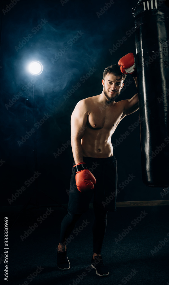 Smiling handsome boxer in red gloves and black sportswear leans against a punching bag and poses for camera. Half-naked man's torso covered in sweat. Concept of strentgh and motivation