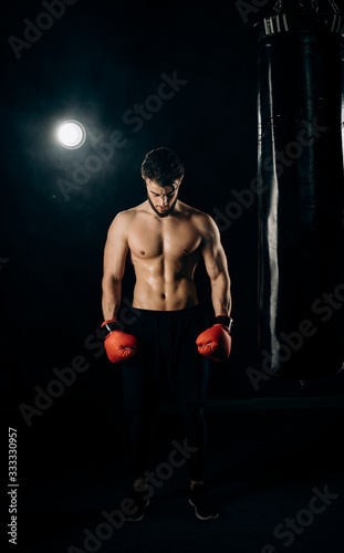 Young athletic boxer in red gloves and black sportswear standing next to a punching bag. Handsome athlete showing muscles and half-naked torso. Concept of strentgh and motivation