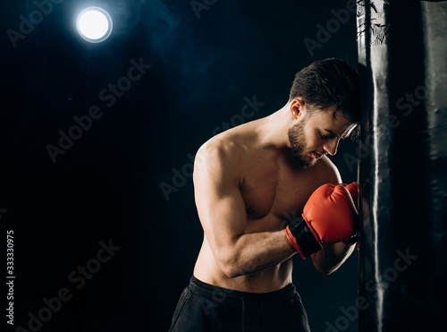 Close up of half naked boxer in red gloves leaning against a punchbag. Young fit boxer during a workout. Athlete punching a punchbag photo