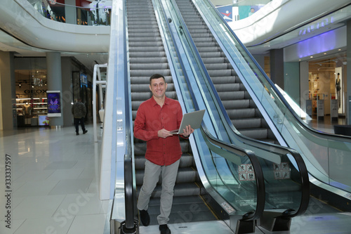 Young handsome businessman in red shirt on an escalator with an open laptop in his hands in a business center of a shopping center or airport.