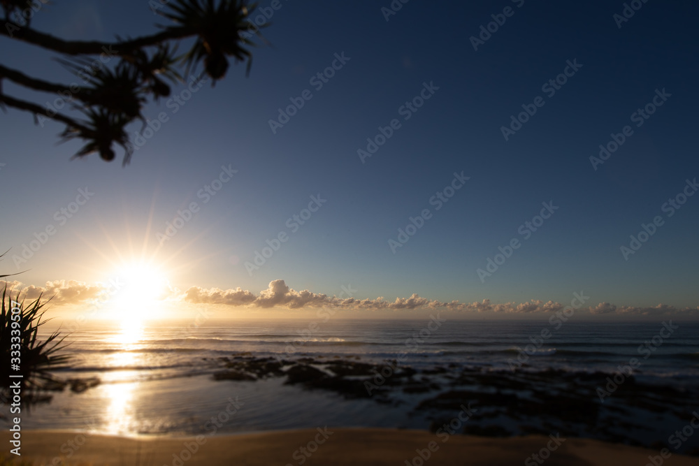 Sun rise at the beach of village of Yeppoon, Queensland, Australia. At the shore of the pacific ocean near Sunshine Coast. Sun rising in between the clouds and reflecting in the water of the sea.  