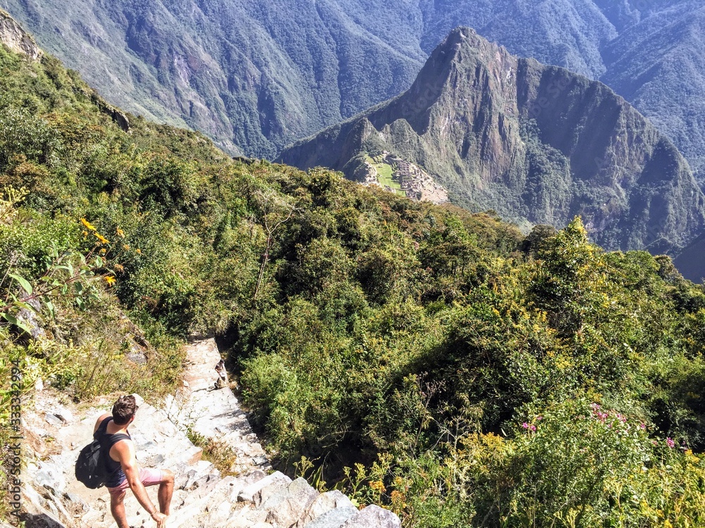 A young male tourist admiring the unique and interesting view of the ancient Inca site of Machu Picchu, nestled high in the Andes Mountains of Peru while climbing Machu Picchi mountain