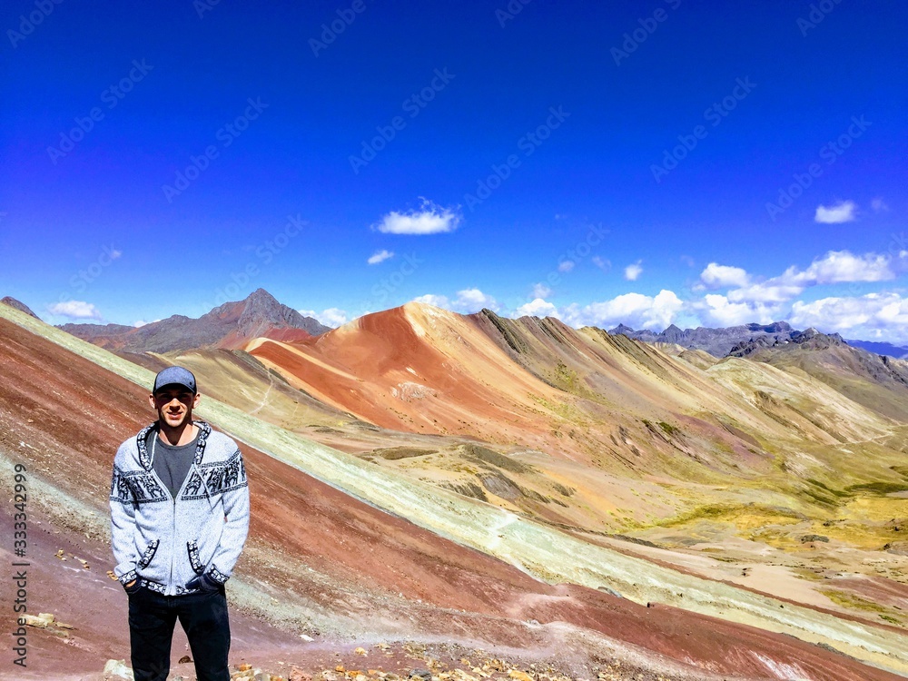 A young male tourist enjoying the view of the incredible Rainbow Mountains outside of Cusco, Peru.  The amounts are a variety of colours from the mineral deposits in the soil.