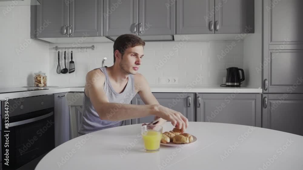 Young man drinking orange juice and using smart device in hand, morning time daily routine of handsome guy sitting at modern kitchen Young man drinking orange juice and using smart device in hand, morning time daily routine of handsome guy sitting at modern kitchen