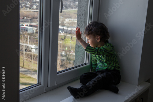 quarantine. a little boy sits in a window-sill and looks out the window bored. longing for fresh air and walks along the street. Forced home during quarantine due to the coronavirus pandemic © tumskaia