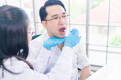 Attractive female doctor taking a saliva sample from asia man mouth with a cotton swab.man chech body during the virus eqidemic