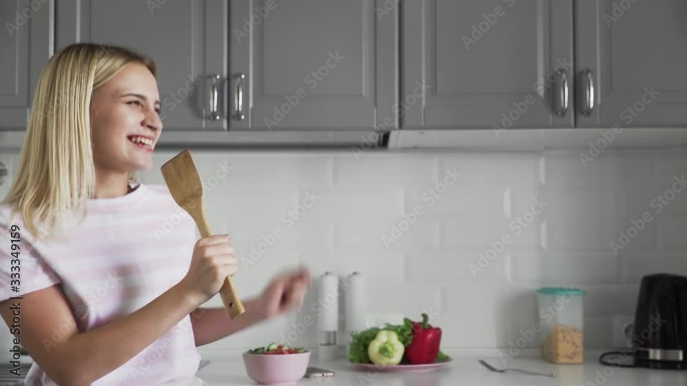 Close up of happy active funny young woman holding wooden spatula microphone singing karaoke song dancing listening music cooking in kitchen Close up of happy active funny young woman holding wooden spatula microphone singing karaoke song dancing listening music cooking in kitchen