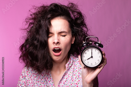  beautiful curly girl in pajamas holds alarm clock in hands, pink background