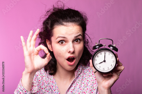  beautiful curly girl in pajamas holds alarm clock in hands, pink background