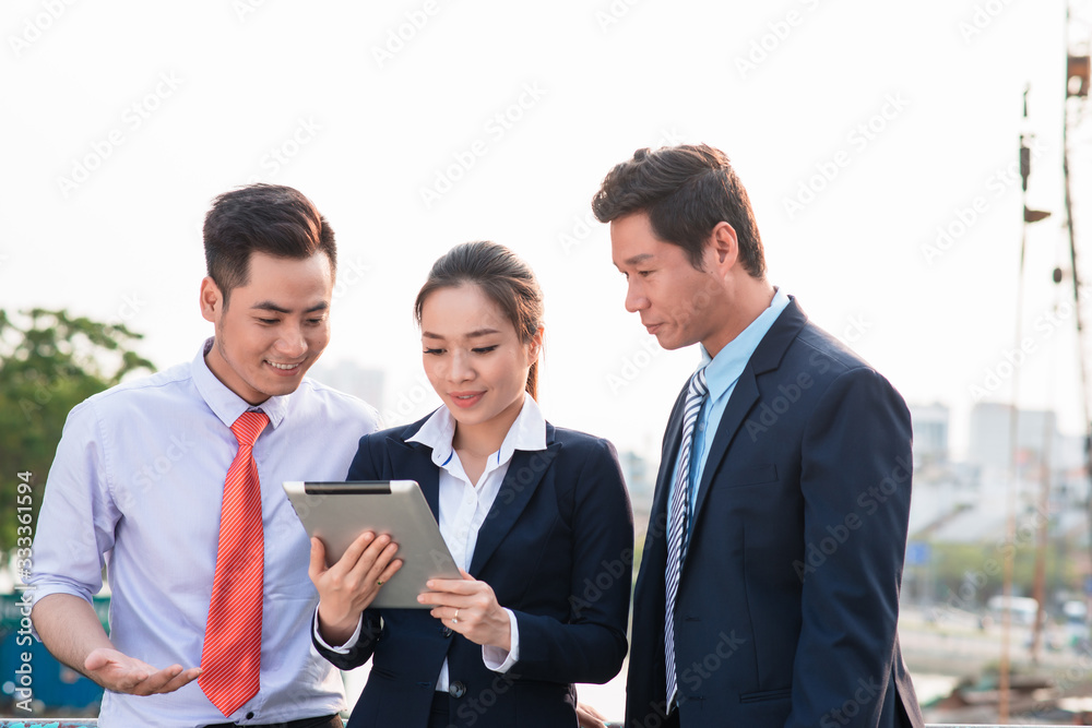 Asian businessman in suit showing online presentation on digital tablet to his colleagues while they standing Asian businessman in suit showing online presentation on digital tablet to his colleagues while they standing
