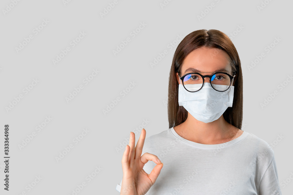 Brunette woman in medical mask and transparent glasses shows ok sign against white background Brunette woman in medical mask and transparent glasses shows ok sign against white background
