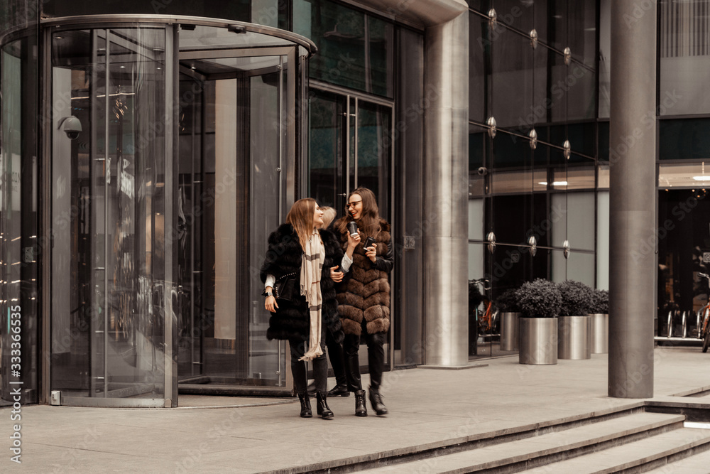 Female friendship, relationships. Two girls are drinking coffee on the street near glass office buildings, a corporation, a bank. The end of the working day of the manager. Coffee on the run.