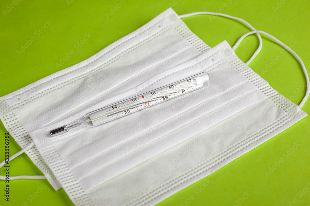 Thermometer and medical mask on a light green background close-up. defence from spread 2019-nCoV, virus outbreak covid-19 quarantine Thermometer and medical mask on a light green background close-up. defence from spread 2019-nCoV, virus outbreak covid-19 quarantine