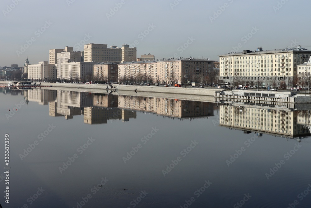 Fototapeta premium Buildings of the Frunze Embankment in Moscow are reflected in the smooth blue water of the Moscow River. Early spring, sunny day.