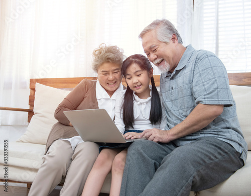 Little granddaughter teach senior elder to surf internet using computer and technology& modern lifestyle.Happy asian grandparent with little young cute grandchild sitting on sofa playing laptop 