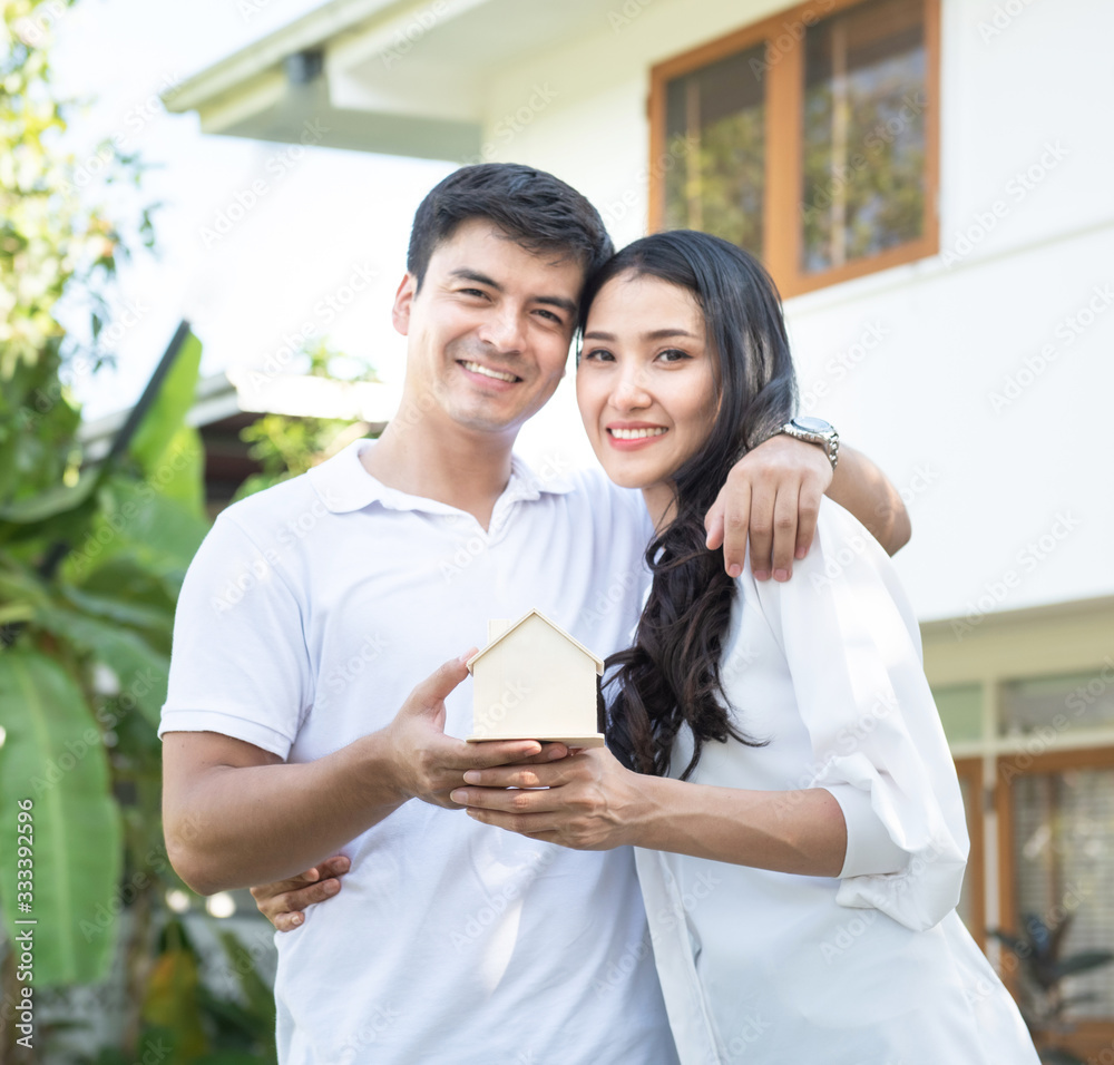 Focused on house wooden shape on hands of asian happy couple standing outside in front of home.Real estate and property housing business concept. Focused on house wooden shape on hands of asian happy couple standing outside in front of home.Real estate and property housing business concept.