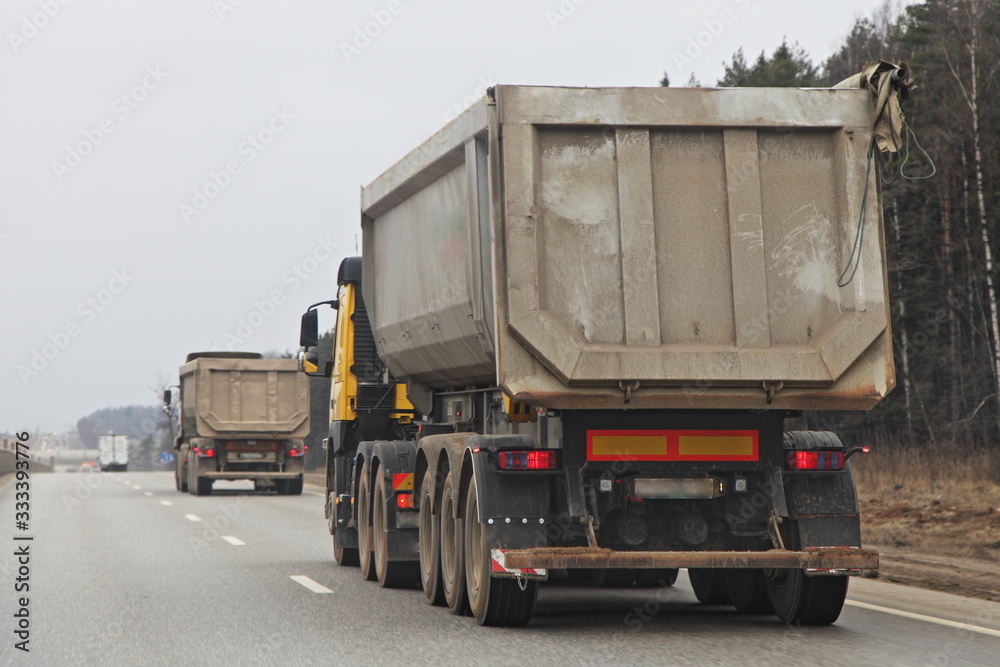 Gray heavy dump truck drive on ground construction site on suburban asphalted highway road at spring day, close up rear-side view – Logistics, bulk cargo transportation trucking