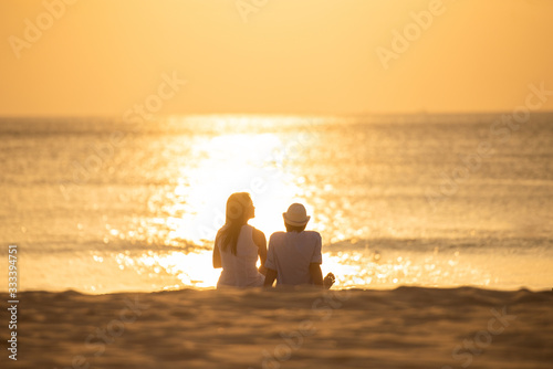 couple sit on sand looking sunset in sea together.