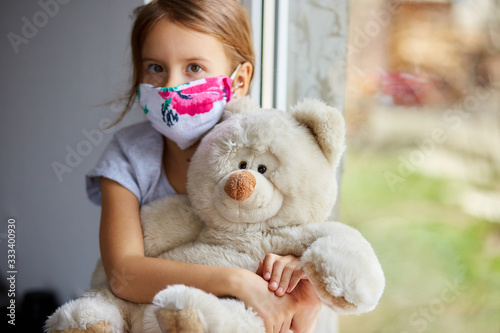Little girl, child in mask with teddy bear sits on windows, coronavirus quarantine