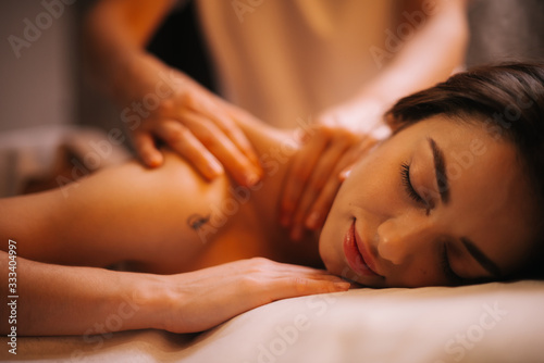 Close-up of face of young relaxed woman lying down on massage table with close eyes who is given back and shoulder massage at spa salon. Concept of luxury massage. Concept of body care.