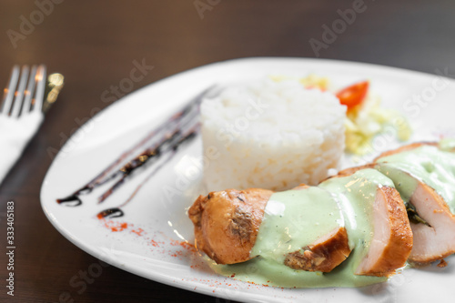 Chicken fillet with sauce is served with rice and vegetables on a white plate and a dark table. Close up.