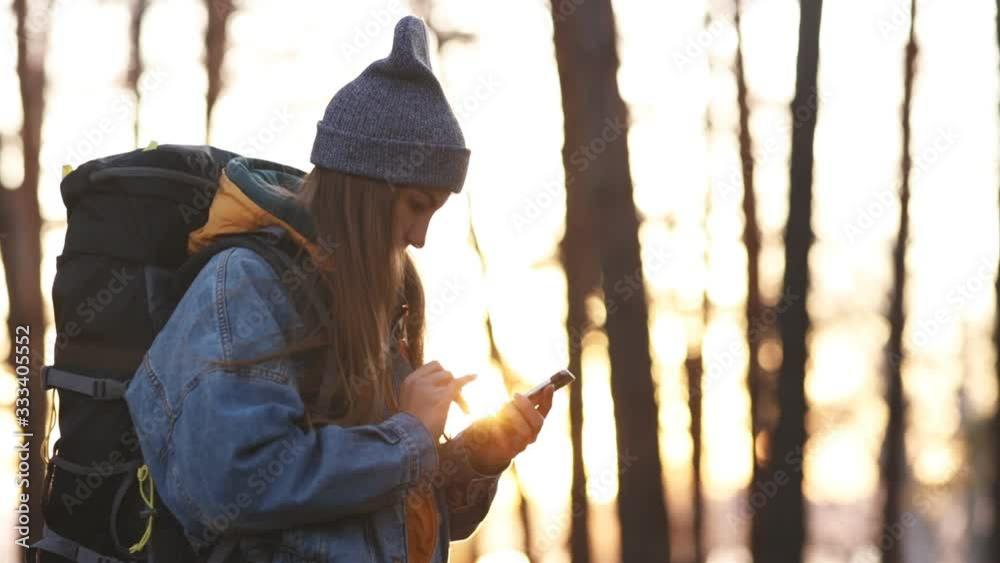 Young charming woman in a denim jacket and a gray hat with a large hiking backpack uses a smartphone in the forest. Search for roads on the map. Message. Communication on-line. Traveler. Sunset light. Young charming woman in a denim jacket and a gray hat with a large hiking backpack uses a smartphone in the forest. Search for roads on the map. Message. Communication on-line. Traveler. Sunset light.