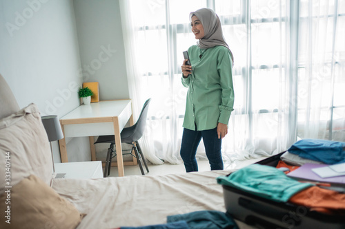 asian muslim woman looking departure schedules via mobile phone after preparing and put her clothes in the suitcase