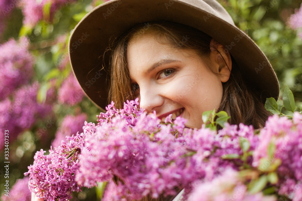 Charming and smiling girl in a hat hides her eyes behind a lush branch of lilac..