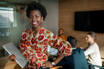 Beautiful young smiling professional black african business woman holding laptop, coworkers hold a meeting in background