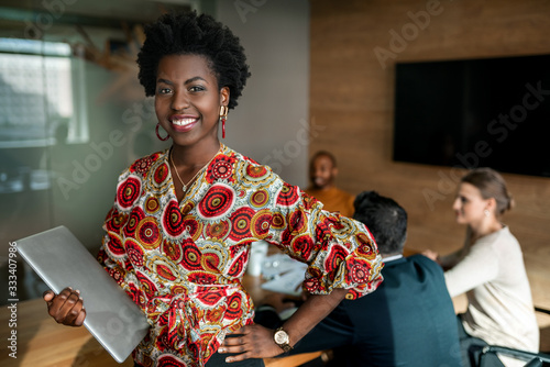 Beautiful young smiling professional black african business woman holding laptop, coworkers hold a meeting in background photo