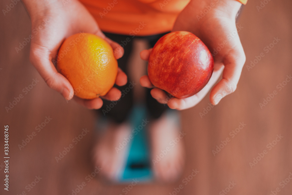 A man in sportswear is standing on the scales and holding in his hands the fruits of an apple and an orange. Weight loss and healthy eating. A man in sportswear is standing on the scales and holding in his hands the fruits of an apple and an orange. Weight loss and healthy eating.