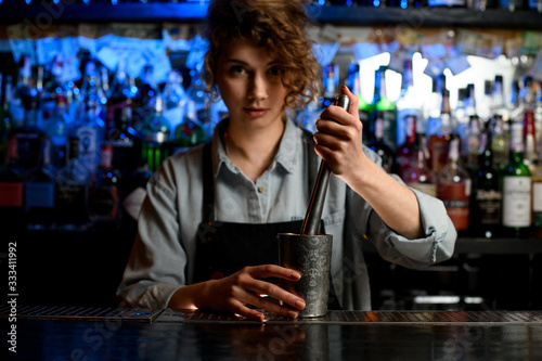 Woman barman in black apron holds steel glass and metal madler.