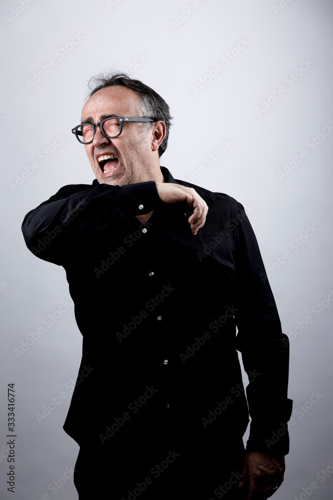 Obraz premium uomo in camicia nera con occhiali starnutisce e si ripara con il gomito, isolato su sfondo nero